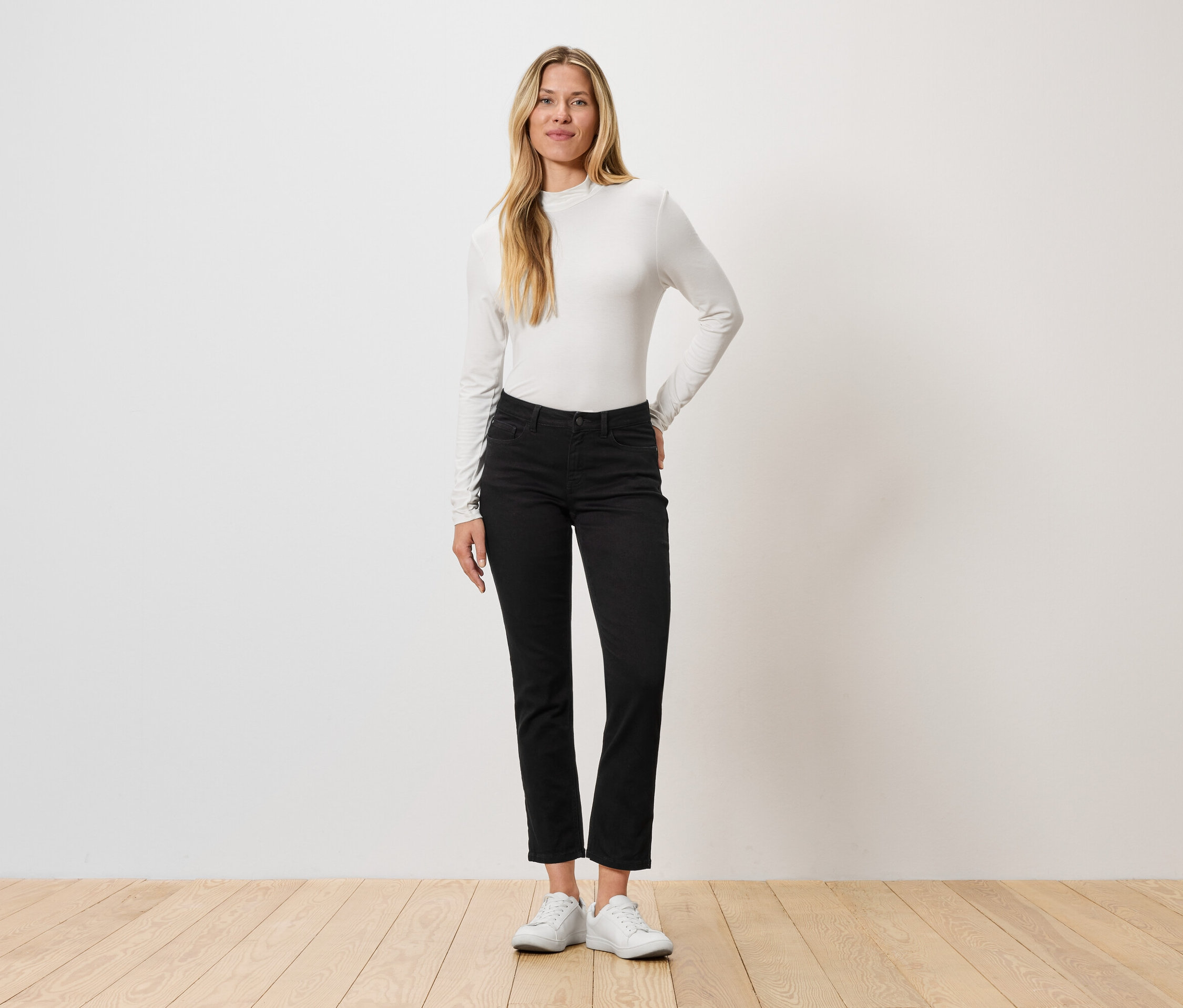 Eine Frau steht vor einer Wand, sie trägt ein langärmliges Oberteil, schwarze Jeans und weiße Turnschuhe.