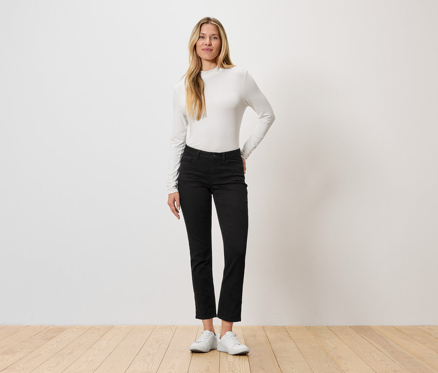 Eine Frau steht vor einer Wand, sie trägt ein langärmliges Oberteil, schwarze Jeans und weiße Turnschuhe.