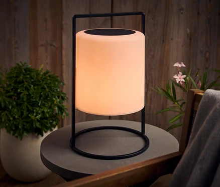 Eine leuchtende Lampe mit schwarzem Gestell steht auf einem grauen Tisch im Freien.