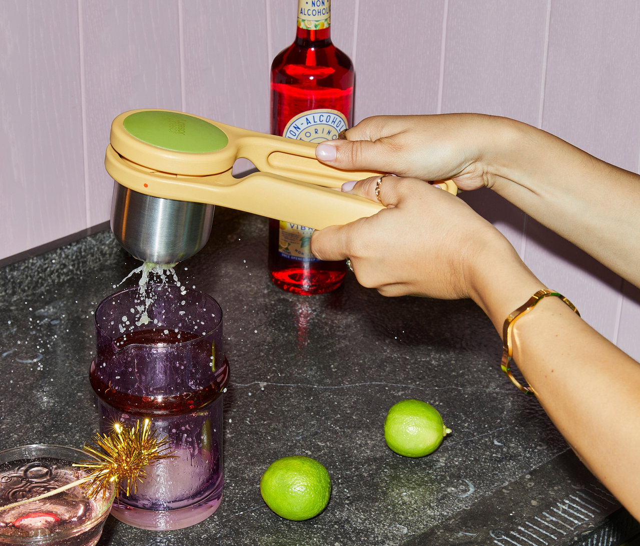Eine Person presst mit einer gelben Zitruspresse Limettensaft in ein lila Glas. Eine Flasche und Limetten liegen auf der Arbeitsfläche.