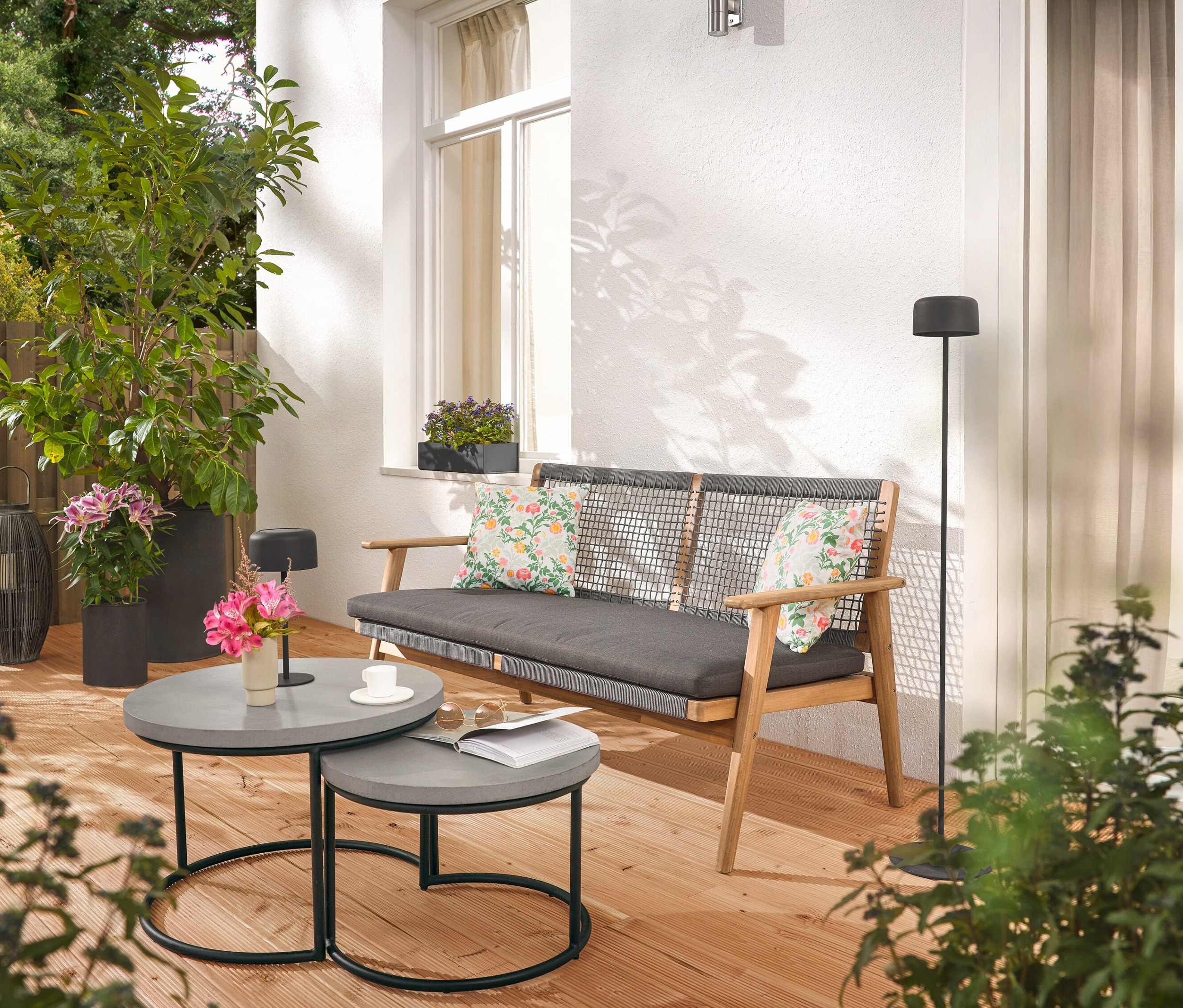 Auf einer Terrasse stehen ein Sofa mit Kissen, zwei graue Tische, eine Stehlampe und Pflanzen.