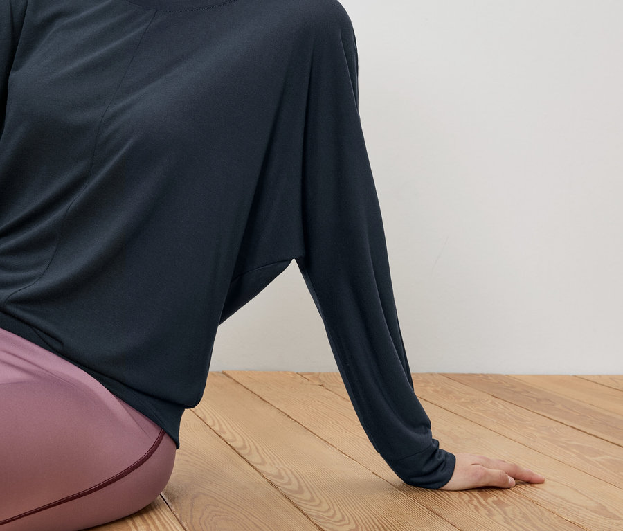 Frau sitzt mit gekreuzten Beinen auf einem Holzboden, trägt ein dunkelblaues Oberteil und rosafarbene Leggings.