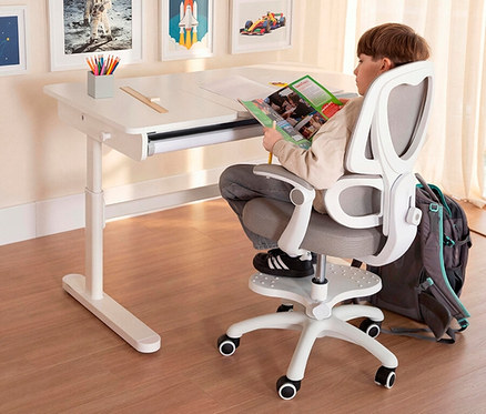 Ein Junge sitzt auf einem grauen Bürostuhl an einem weißen Schreibtisch und liest ein Magazin. Ein grauer Rucksack steht neben dem Stuhl.