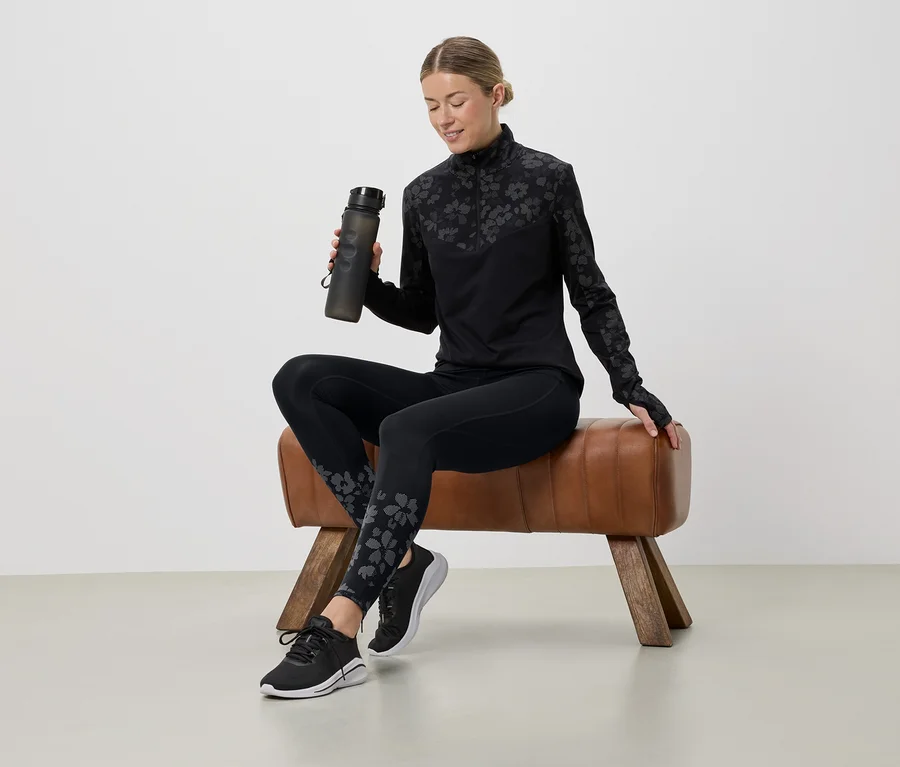 Frau sitzt auf einem Turnbock, hält eine Wasserflasche und trägt ein schwarzes Sportoutfit mit Blumenmuster.