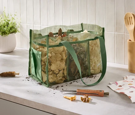 Auf einer Küchentheke steht eine grüne Netztasche voller Pilze neben einem Messer, einem Küchentuch mit Pilzaufdruck und einem Topf mit Kräutern.