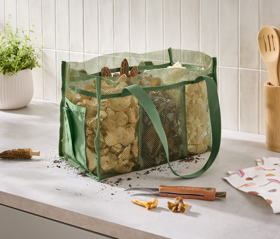 Auf einer Küchentheke steht eine grüne Netztasche voller Pilze neben einem Messer, einem Küchentuch mit Pilzaufdruck und einem Topf mit Kräutern.