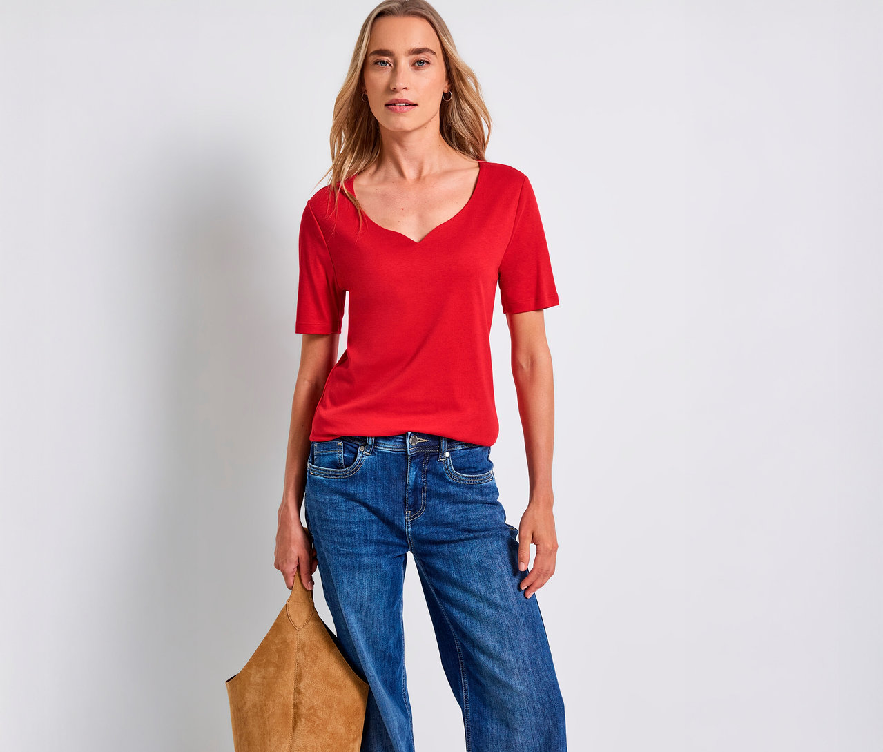 Frau trägt ein rotes Street One Shirt mit Herzausschnitt, blaue Jeans und hält eine braune Tasche.