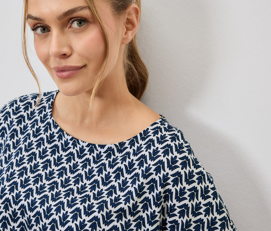 Porträt einer Frau mit blonden Haaren, die ein blau-weißes Oberteil mit Blattmuster trägt.