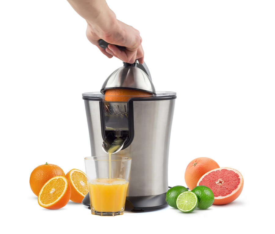 Eine Hand drückt eine Orange in eine silberne Saftpresse, Saft läuft in ein Glas, umgeben von Orangen, Limetten und Grapefruit.