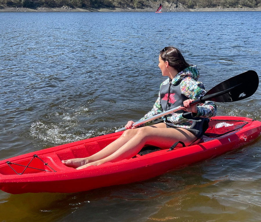 Frau paddelt in einem roten Kajak auf einem See. Sie trägt eine gemusterte Jacke und eine Schwimmweste.