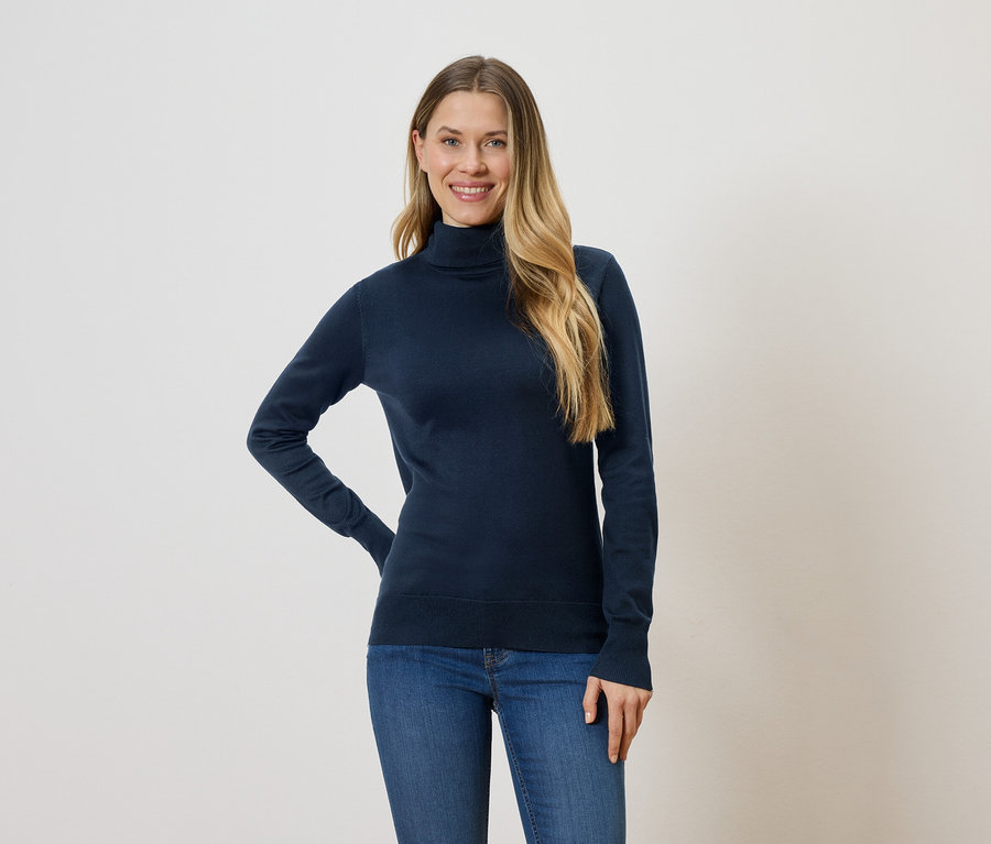 Frau mit langen blonden Haaren, die einen marineblauen Rollkragenpullover und Jeans vor einem weißen Hintergrund trägt.