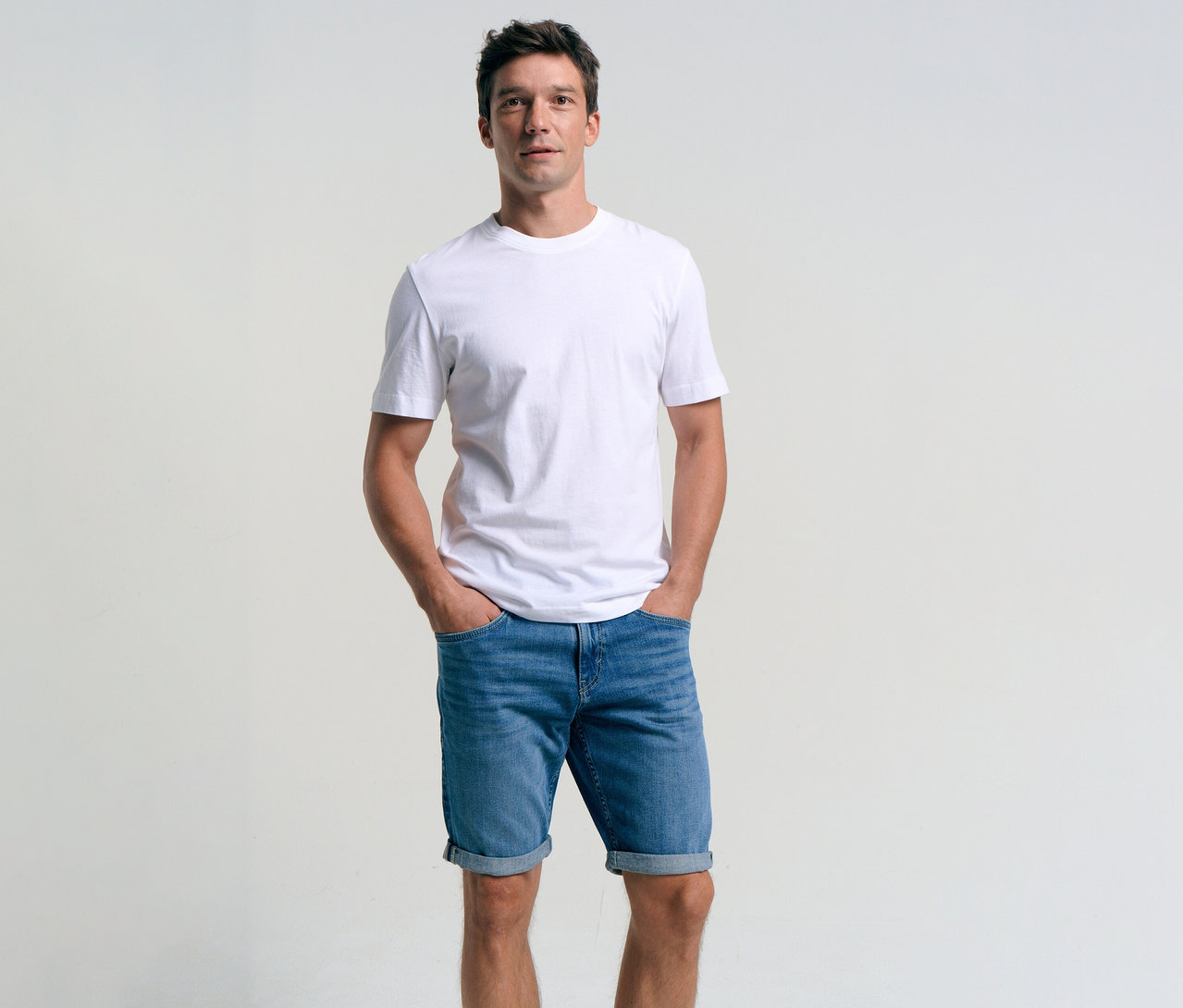 Mann mit weißem T-Shirt und Tom Tailor TTJOSH REGULAR SLIM Jeansshorts in Mid Stone Bright Blue Denim mit Händen in den Hosentaschen.