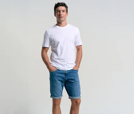 Mann mit weißem T-Shirt und Tom Tailor TTJOSH REGULAR SLIM Jeansshorts in Mid Stone Bright Blue Denim mit Händen in den Hosentaschen.
