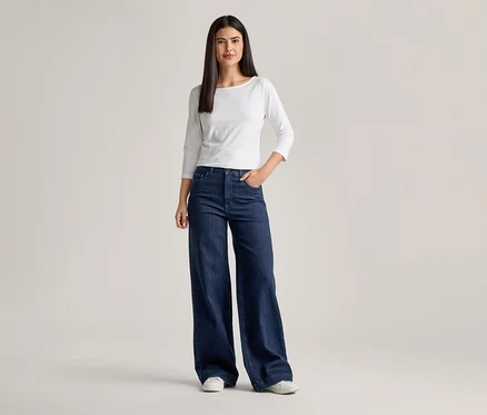 Frau mit weißem Langarmshirt und ausgestellter, dunkelblauer Jeans mit einer Hand in der Hosentasche.