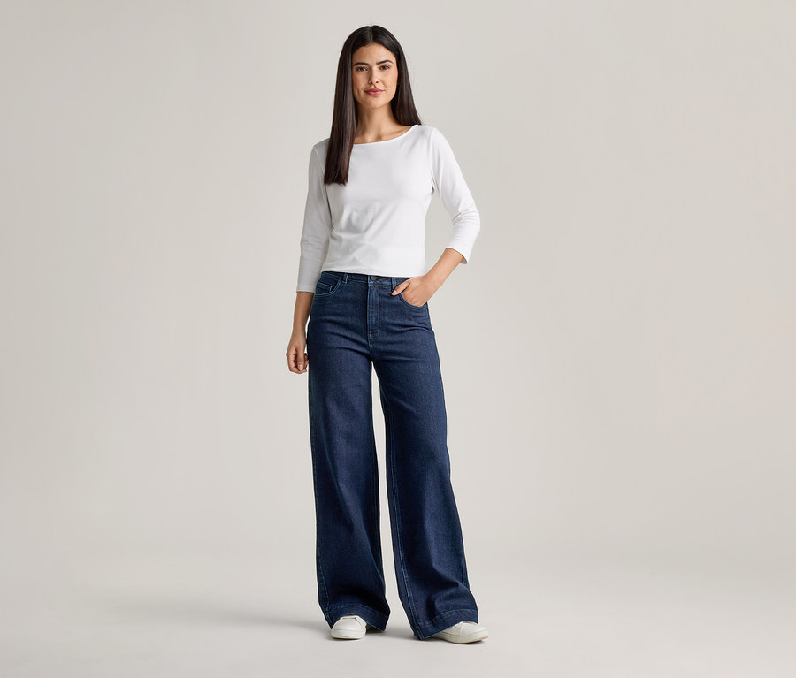 Frau mit weißem Langarmshirt und ausgestellter, dunkelblauer Jeans mit einer Hand in der Hosentasche.