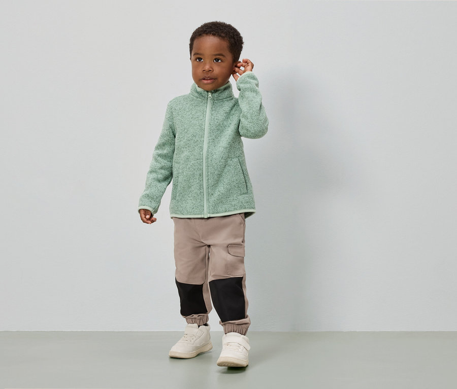 Ein Junge steht vor einer weißen Wand und trägt eine hellgrüne Fleecejacke, beige Hosen mit schwarzen Einsätzen und weiße Schuhe.