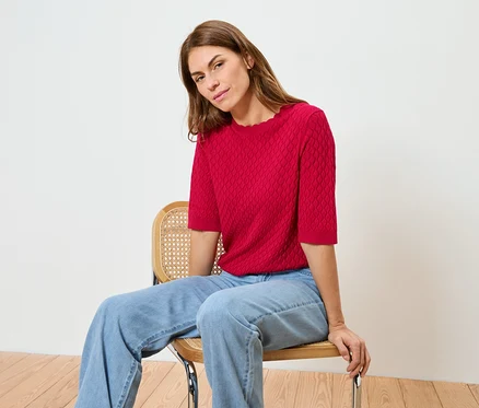 Eine Frau sitzt auf einem Stuhl und trägt ein rotes Oberteil und Jeans.