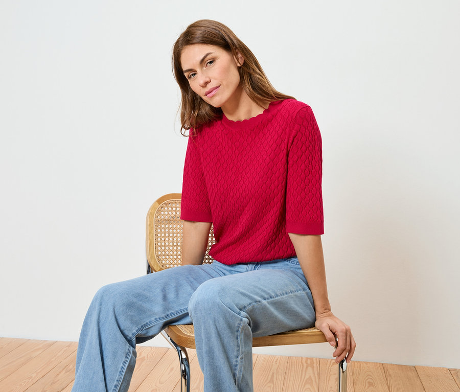 Eine Frau sitzt auf einem Stuhl und trägt ein rotes Oberteil und Jeans.