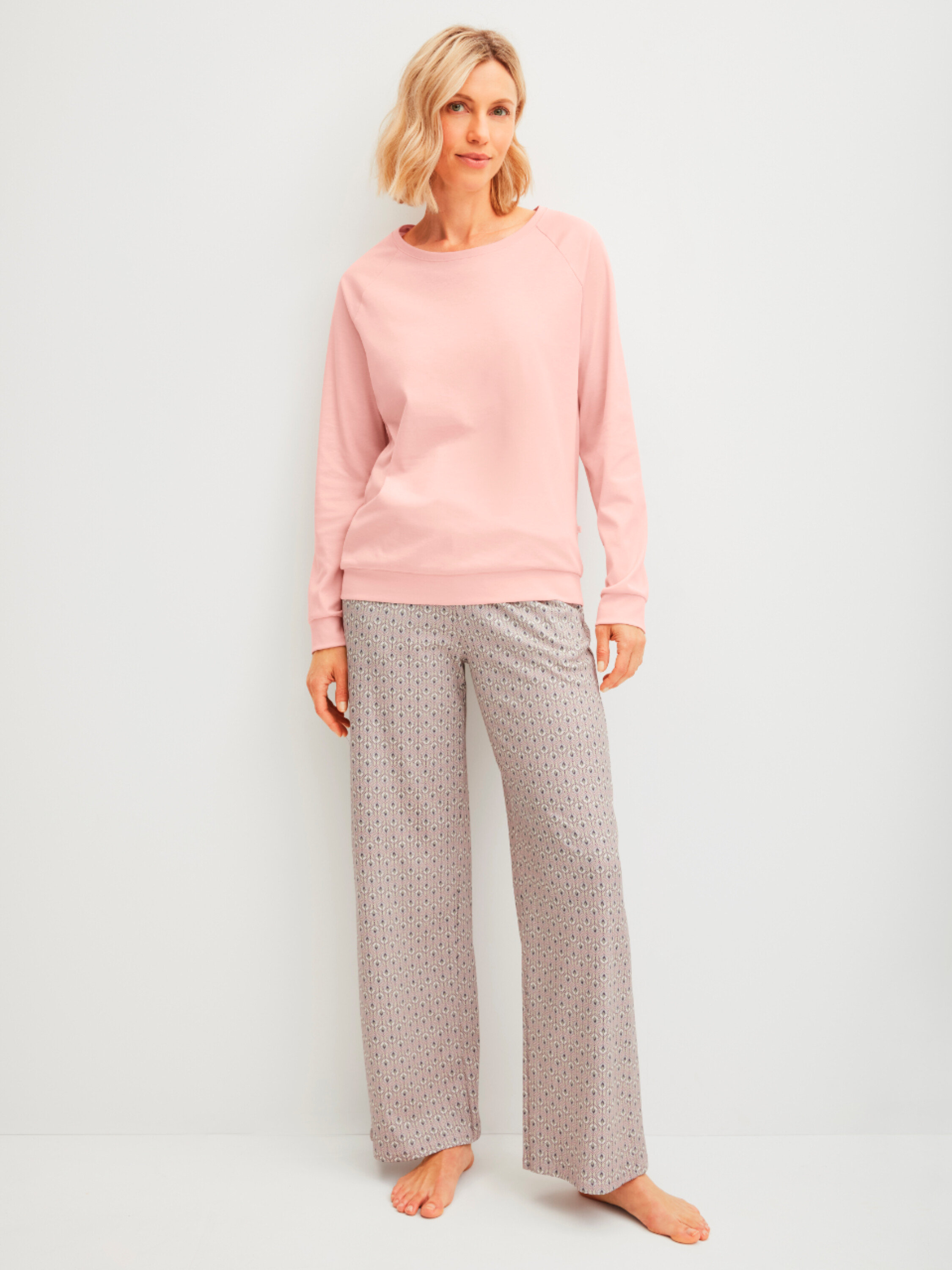 Frau steht barfuß in einem rosafarbenen Pullover und einer gemusterten Hose vor einer weißen Wand.