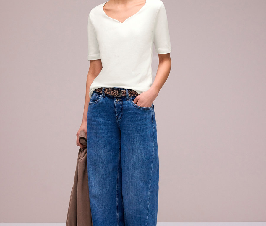 Frau mit lockigem Haar trägt ein weißes Street One Kurzarmshirt, weite blaue Jeans mit Leopardenmustergürtel und helle Flats, eine braune Jacke haltend.