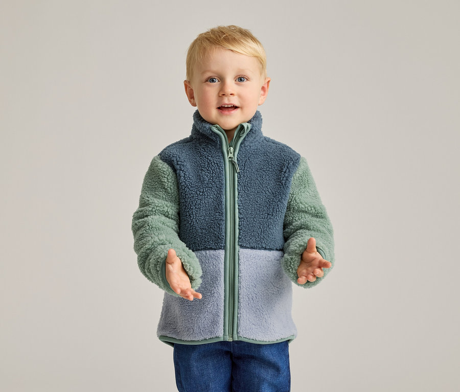 Ein blonder Junge mit blauen Augen trägt eine Fleecejacke in Blau, Grün und Hellblau und blaue Jeans.