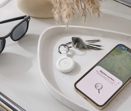 Auf einer Ablage liegen eine Sonnenbrille, Schlüssel, ein weißer Tracker und ein Smartphone mit Karten-App.