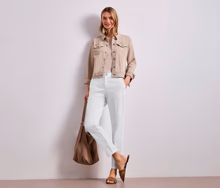 Eine lächelnde Frau steht vor einer weißen Wand und trägt eine beigefarbene Jeansjacke, ein helles Oberteil und eine weiße Street One Chino Hose. Sie hält eine braune Umhängetasche und trägt braune Sandalen.