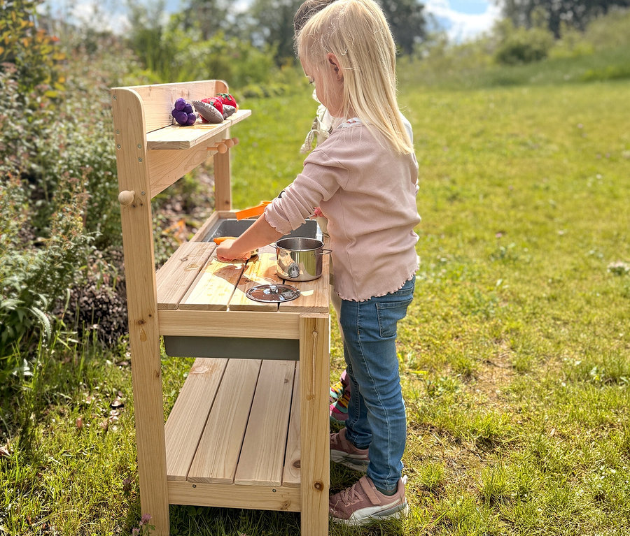 Ein blondes Kind spielt draußen an einer hölzernen Matschküche, hält eine Spielzeugkarotte über einen Topf. Auf dem oberen Regal liegen Spielzeuggemüse.