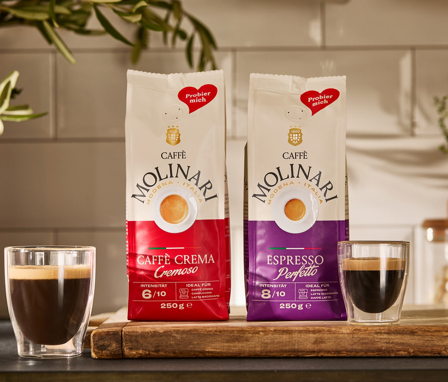 Zwei Packungen Caffè Molinari Caffè Crema & Espresso und zwei Tassen Kaffee auf einem Holzbrett.
