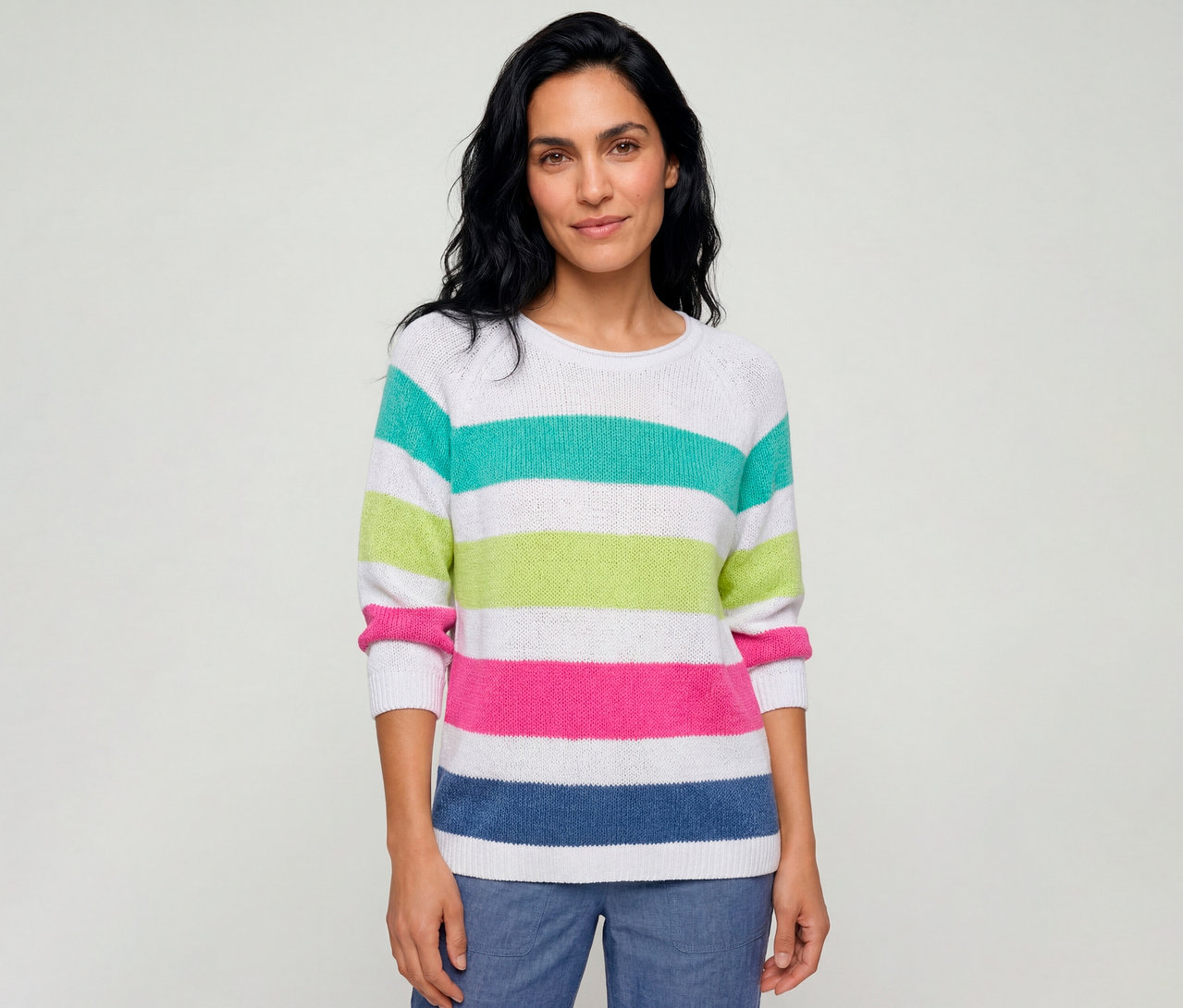 Eine Frau trägt einen gestreiften Olsen Pullover in Weiß mit türkisen, lindgrünen, pinken und blauen Streifen und blaue Hosen.