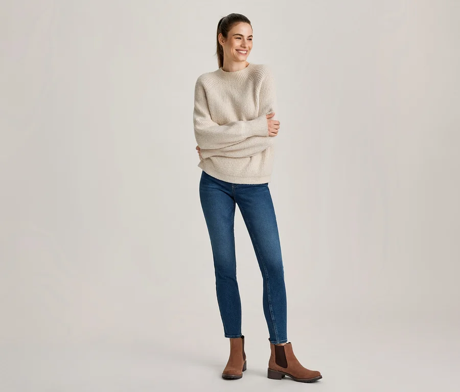 Frau mit Pferdeschwanz, lächelnd und nach rechts blickend, trägt einen cremefarbenen Rippstrickpullover, blaue Skinny Jeans und braune Stiefeletten.