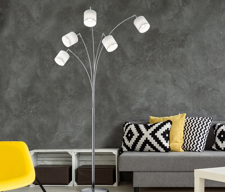 Ein modernes Wohnzimmer mit einer silbernen Stehlampe mit fünf Lichtern. Rechts steht ein graues Sofa mit schwarz-weiß-gelben geometrischen Kissen, links ein gelber Stuhl. Ein schwarz-weißer geometrischer Teppich bedeckt den Boden vor einer dunkelgrauen strukturierten Wand.