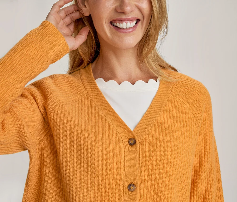 Eine lächelnde Frau in einem orangefarbenen Rippstrick-Cardigan über einem weißen Oberteil mit Wellenkante, eine Hand am Ohr.