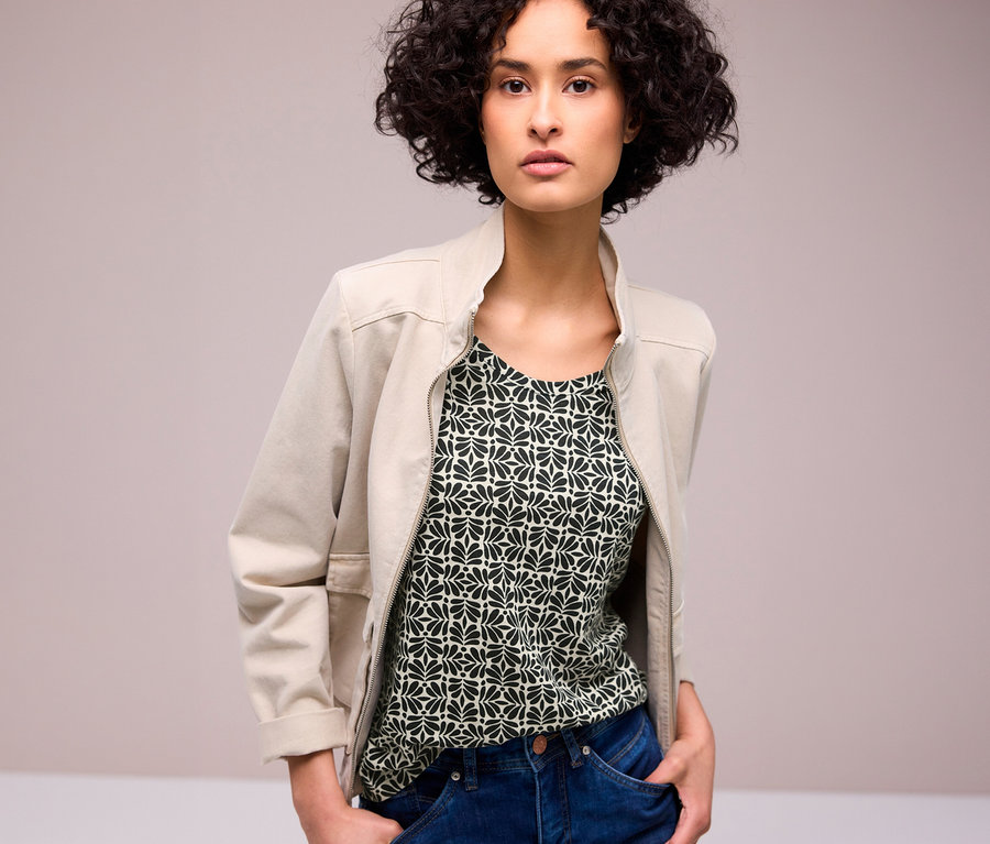 Eine Frau trägt eine offene beige Jacke über einem gemusterten Street One T-Shirt und blauen Jeans. Ihre Hände stecken in den Taschen.