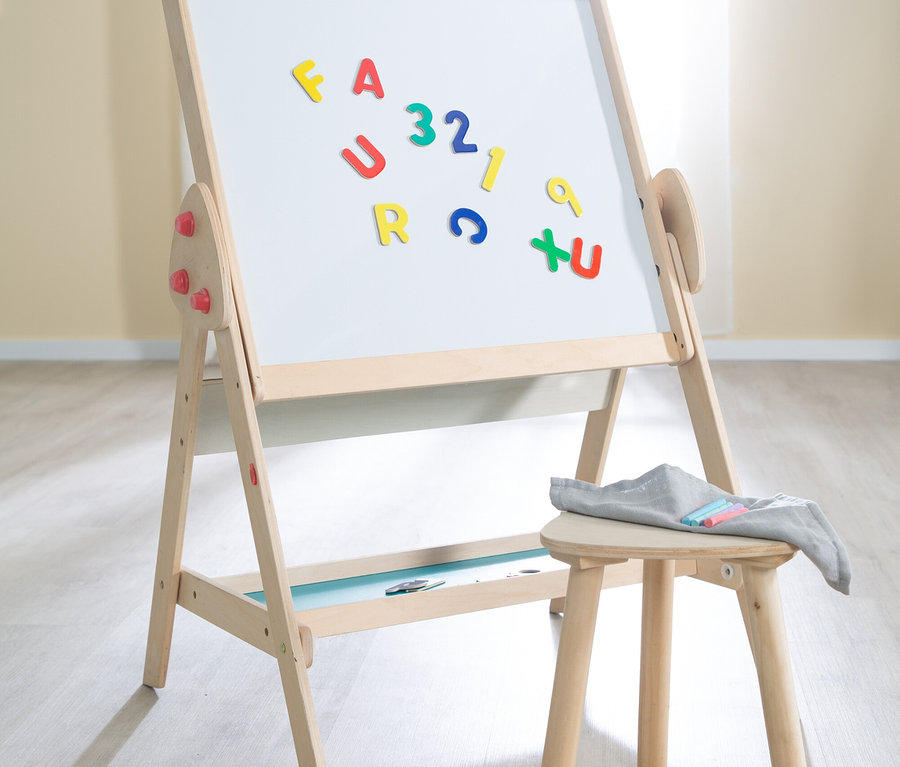 Ein hölzerner Staffelei mit einer weißen Tafel steht auf einem hellen Holzboden. Bunte Magnetbuchstaben und -zahlen sind auf der weißen Tafel angeordnet. Rechts daneben steht ein kleiner Holzschemel mit einem grauen Tuch und mehreren farbigen Kreidestücken.