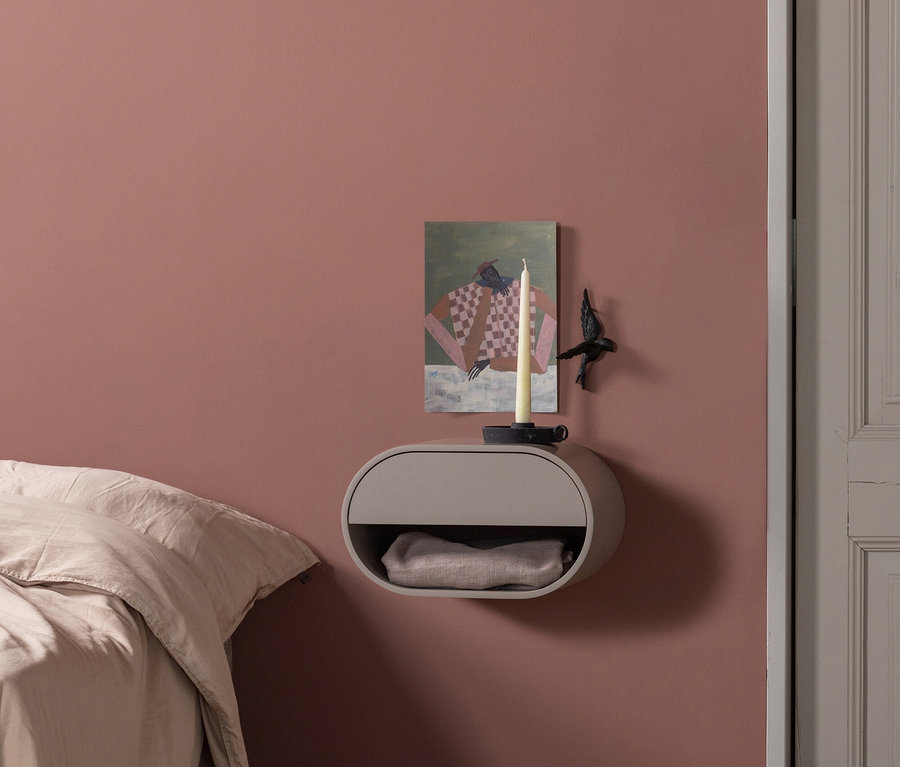 Schlafzimmer mit pink-roter Wand, links ein Bett mit heller Bettwäsche. In der Mitte ein grauer, ovaler Wand-Nachttisch mit einem Bild, einer weißen Kerze und einer schwarzen Vogelfigur. Im Fach liegt eine helle Decke. Rechts ein grauer Türrahmen, der Boden ist heller Holzboden im Fischgrätmuster.