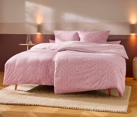 Ein Bett mit rosa gemusterten Bettbezügen und Kissen sowie unifarbenen rosa Laken steht auf einem hellen Teppich auf Holzboden. Links daneben ein kleiner runder Tisch mit Wasserflasche und Glas. Zwei Wandleuchten erhellen die zweifarbige rosa Wand hinter dem Bett, rechts steht eine hohe Vase.