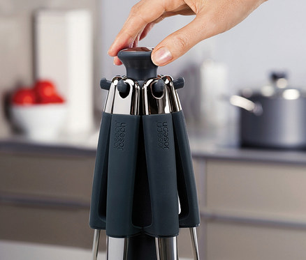 Eine Hand drückt auf die Oberseite eines dunkelgrauen und silbernen Joseph Joseph Elevate Steel Carousel Küchenutensilienhalters vor unscharfem Küchenhintergrund.