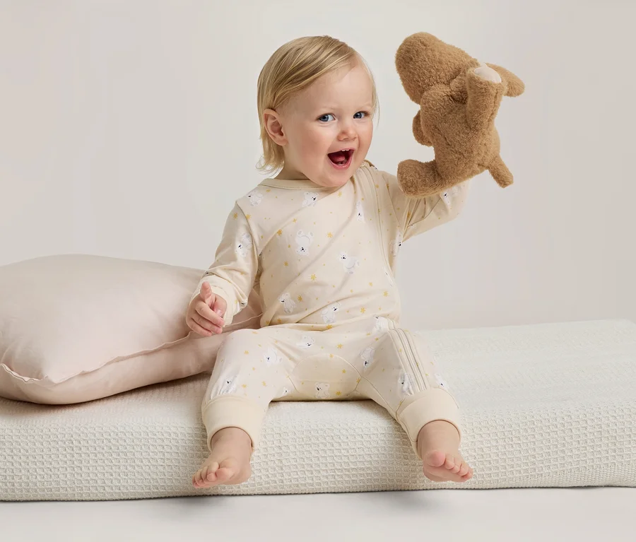 Ein lächelndes Baby in hellgelbem Schlafanzug mit Schafmuster sitzt auf einer weißen Decke und hält ein braunes Stofftier hoch. Daneben liegt ein rosa Kissen.