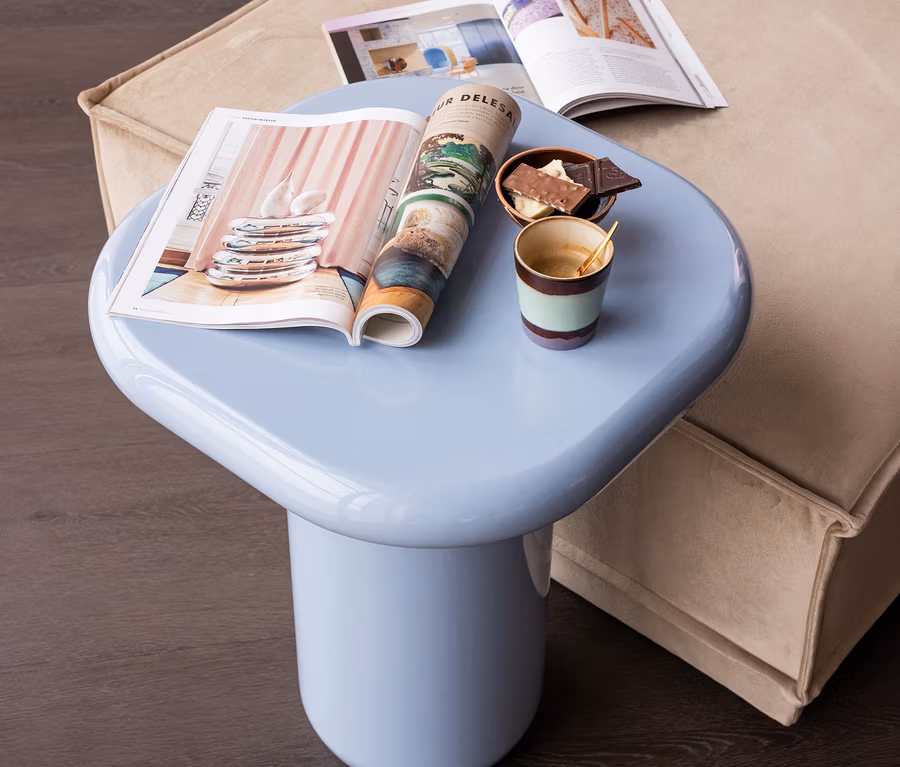 Ein hellblauer Beistelltisch mit unregelmäßiger Platte, darauf zwei offene Zeitschriften, eine Schale mit dunkler und weißer Schokolade und eine Tasse Kaffee mit Löffel, steht neben einem beigen Sofa.