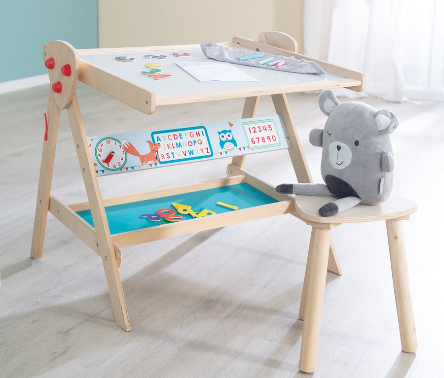 Ein hölzerner Kindermaltisch mit weißer Tafel, einer bunten Lernleiste und einer blauen Ablage. Magnetbuchstaben und eine Zeichnung liegen auf der Tafel, Buntstifte in einem Beutel. Ein grauer Plüschbär sitzt auf einem kleinen Holzhocker neben dem Maltisch.