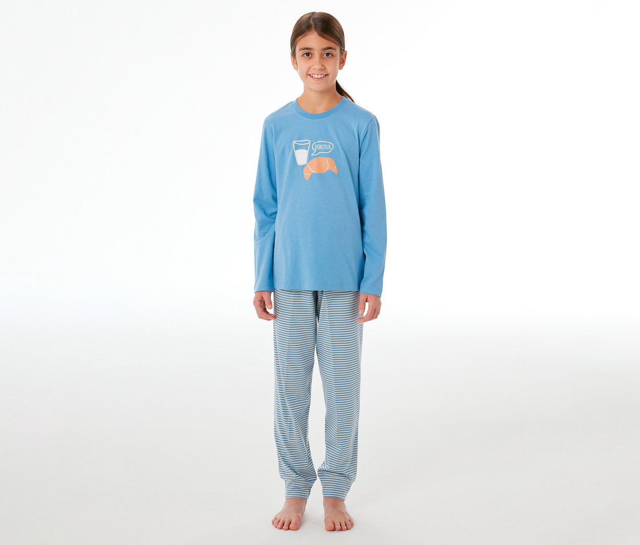 Lächelndes Mädchen in einem SCHIESSER Kinder-Pyjama. Das hellblaue Oberteil hat ein Glas Milch, ein Croissant und "Bonjour" aufgedruckt. Die Hose ist hellblau-weiß gestreift.