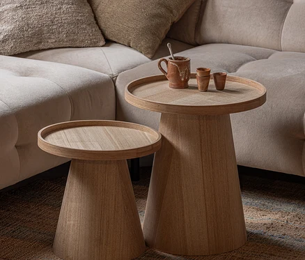 Zwei helle Holztische mit runden Platten und konischen Füßen, einer höher, stehen neben einem beigen Sofa. Auf dem höheren Tisch stehen ein rustikaler Becher und zwei kleine Tassen. Sie stehen auf einem strukturierten Teppich.