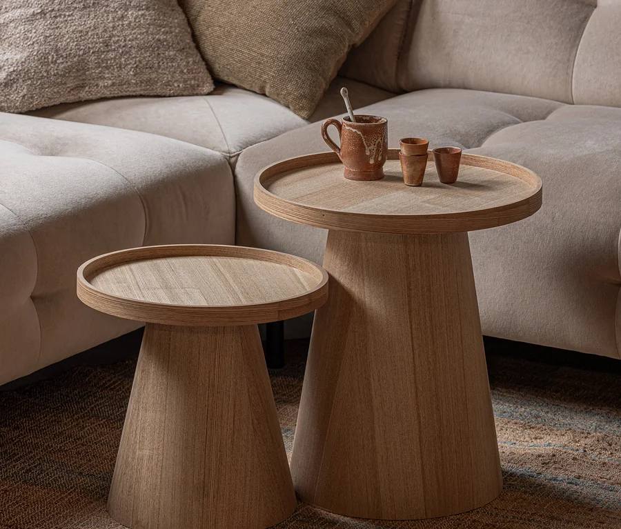 Zwei helle Holztische mit runden Platten und konischen Füßen, einer höher, stehen neben einem beigen Sofa. Auf dem höheren Tisch stehen ein rustikaler Becher und zwei kleine Tassen. Sie stehen auf einem strukturierten Teppich.