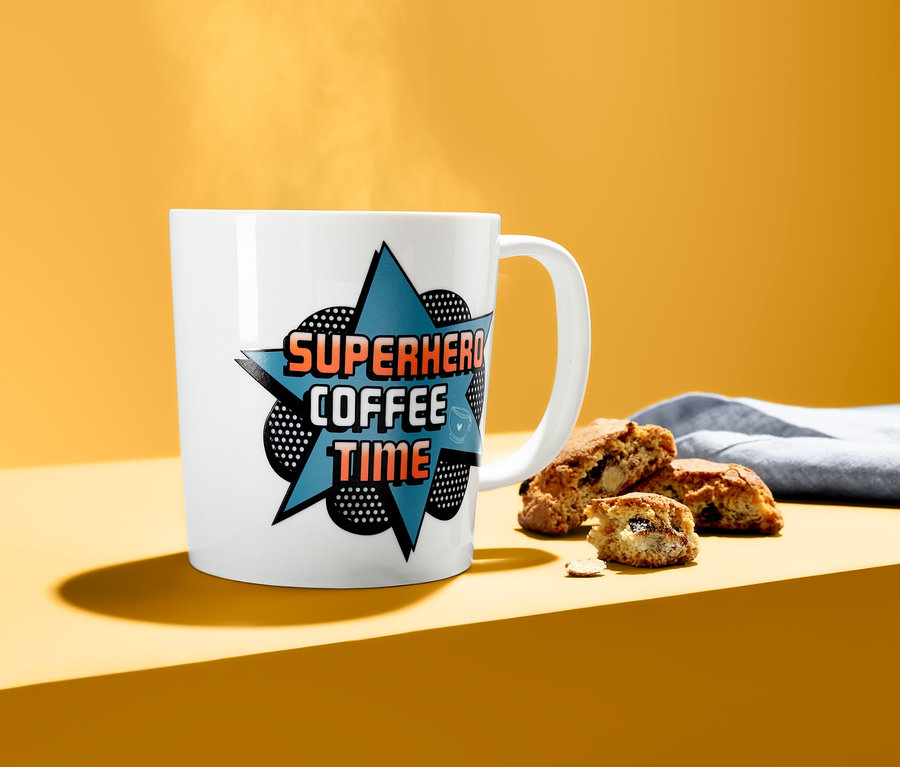 Weißer Trinkbecher mit Farbwechseleffekt mit dem Aufdruck SUPERHERO COFFEE TIME neben Keksen.
