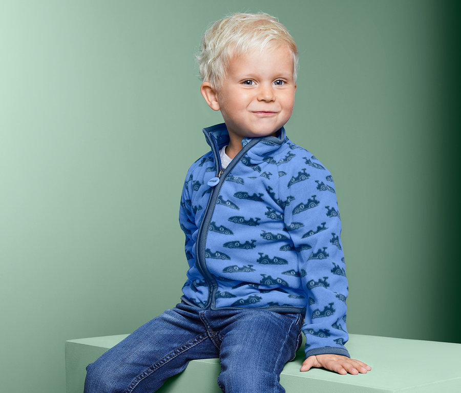 Junge sitzt auf einem Block und trägt eine blaue Microfleecejacke mit Aufdruck.