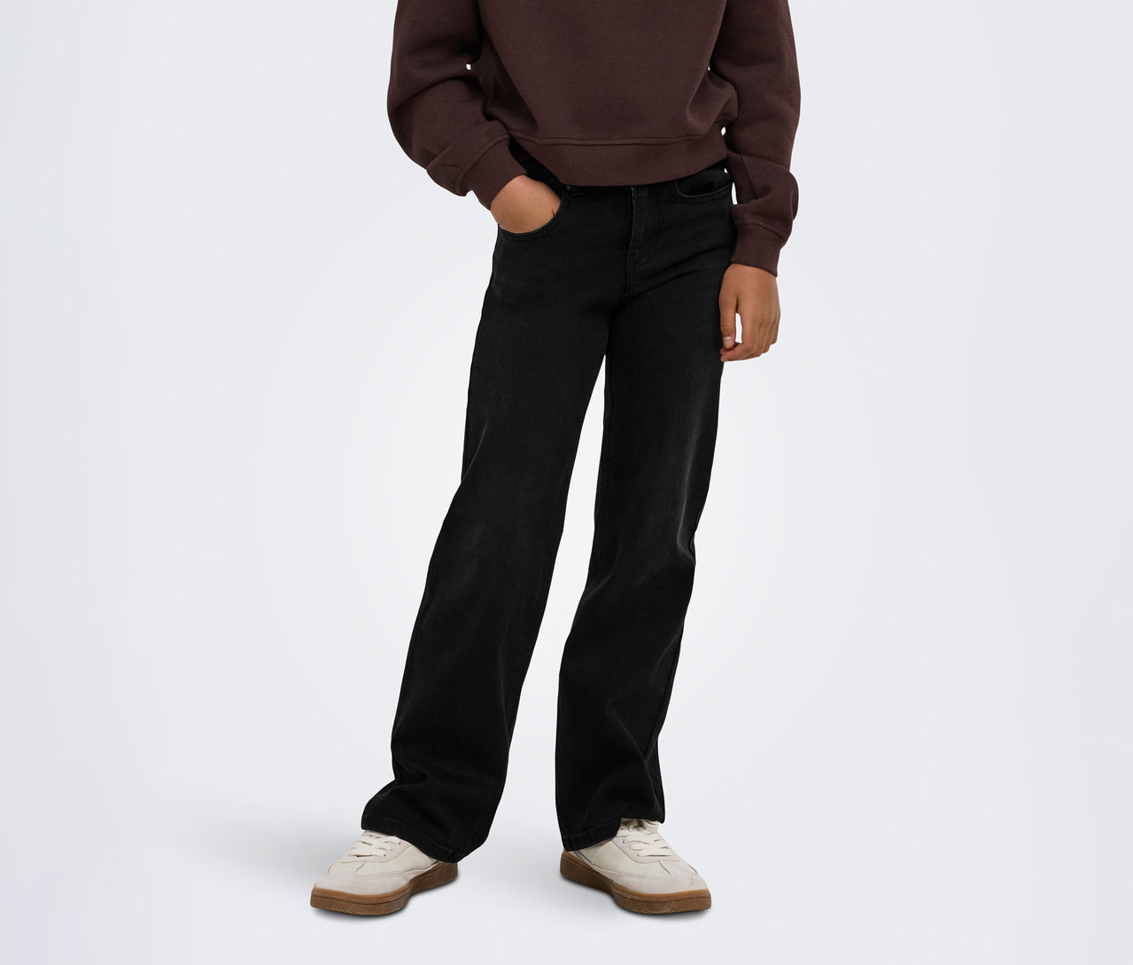 Person trägt ein braunes Sweatshirt, schwarze weite Jeans mit einer Hand in der Tasche und hellbeige Sneaker.