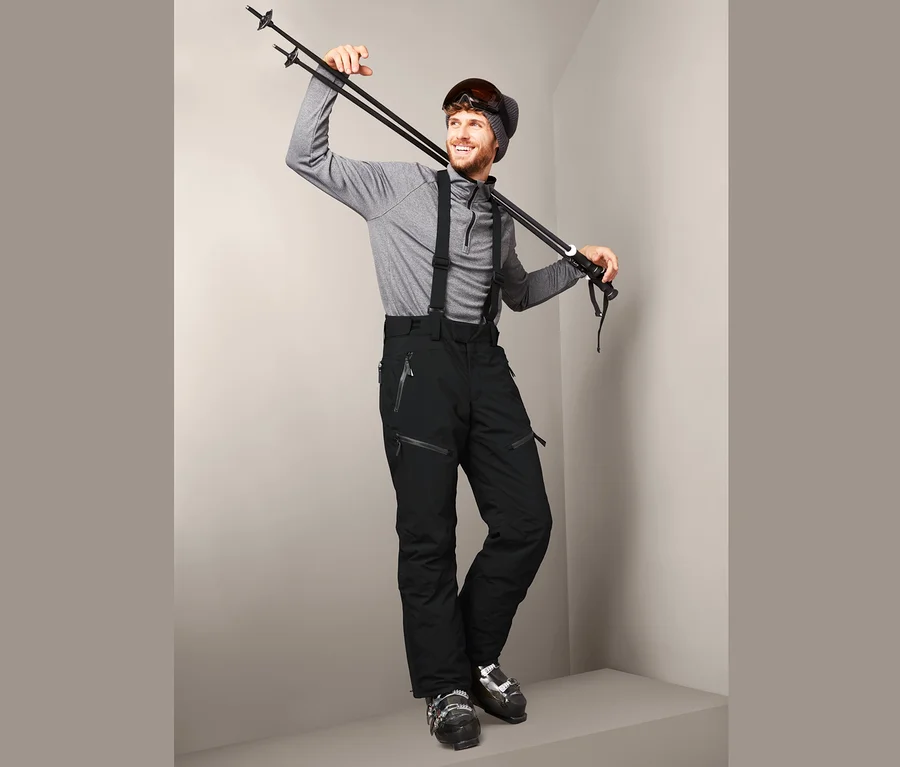 Mann posiert mit Skistöcken über der Schulter, trägt eine schwarze High-End-Skihose und Skischuhe.