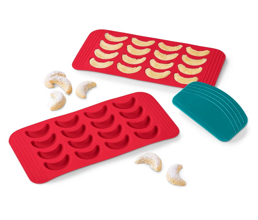 Zwei rote Vanillekipferl-Backmatten, eine mit gebackenen Kipferln, einige Kipferl daneben und ein türkisfarbener Schaber.