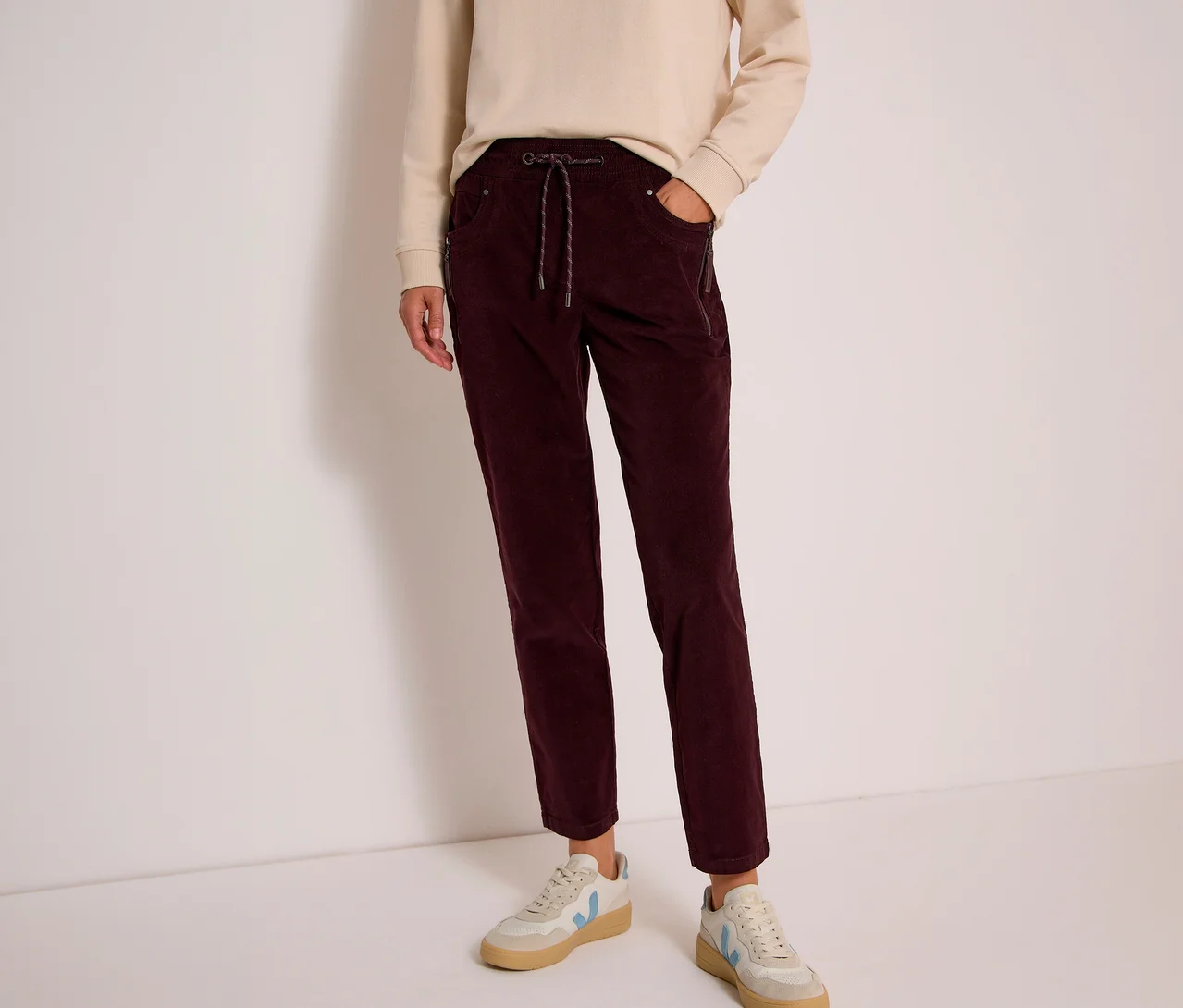 Person trägt ein beigefarbenes Sweatshirt und eine dunkelrote CECIL Cordhose mit Kordelzug. Dazu helle Sneaker.